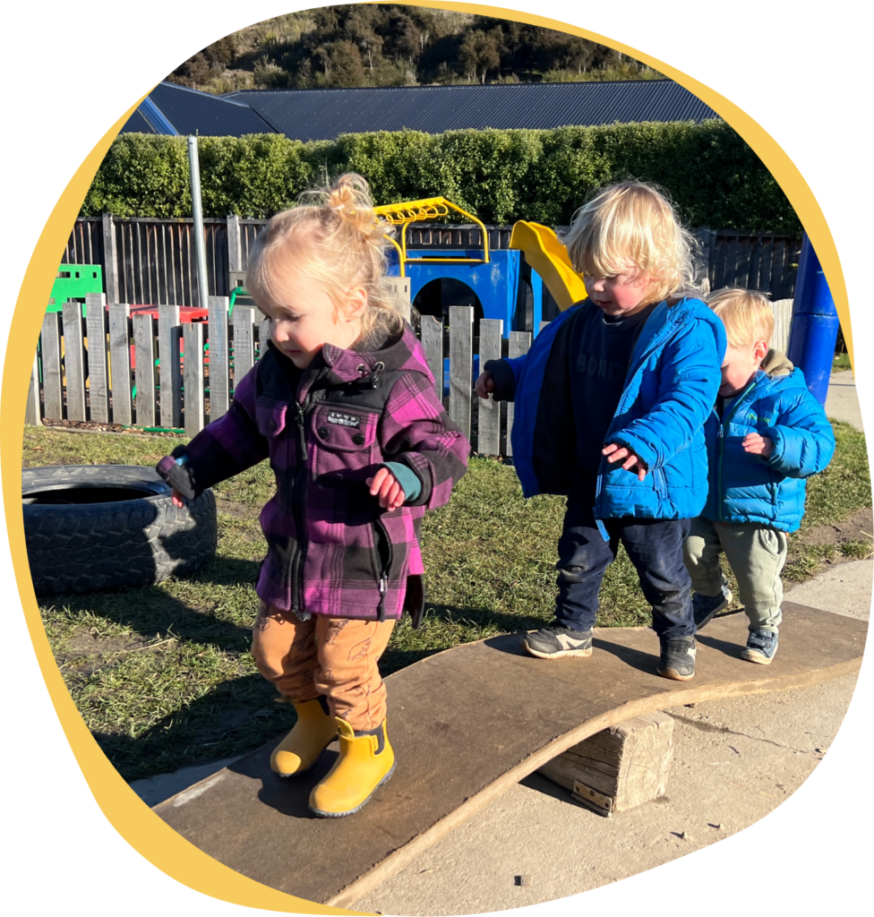 Riverside Early Childcare Three young children enjoying early childcare in Wanaka, playing outdoors at a playground, two of them wearing blue jackets and one in a purple plaid jacket with yellow boots, with toy trucks and wooden blocks
