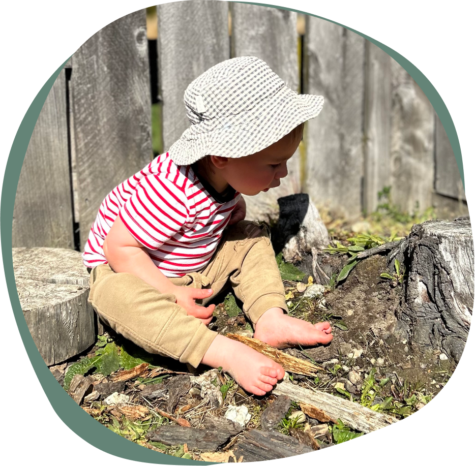 Riverside Early Childcare A toddler in a striped red and white shirt and khaki pants sits on the ground at an early childcare in Wanaka and plays with sticks, wearing a white floppy hat, with a wooden fence and