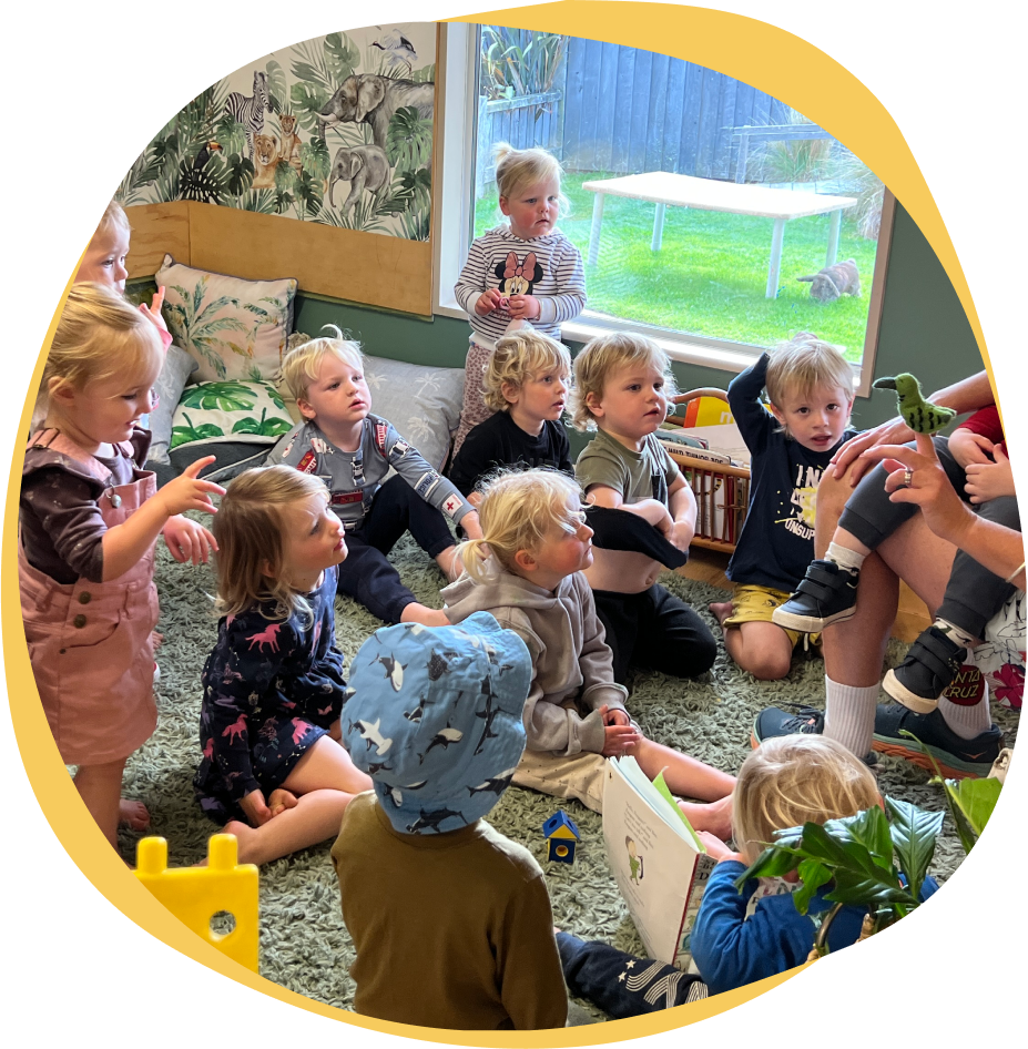 Riverside Early Childcare A group of young children sitting on the floor in an early childcare Wanaka classroom, attentively listening to an adult who is reading a book and showing them a toy dinosaur. The room is colorful with